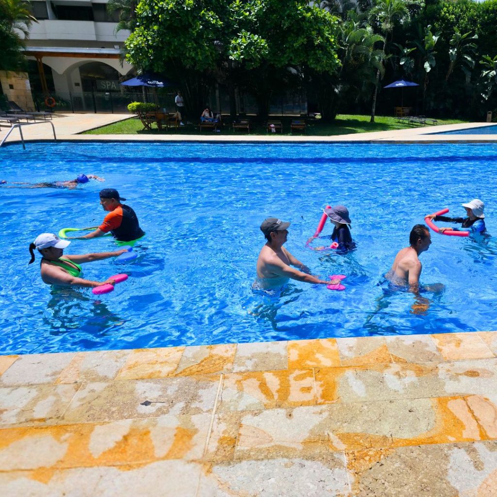 Grupo de hombres y mujeres en una clase dinámica de AquaFitness usando flotadores tubulares en la piscina del Hotel Intercontinental Cali. Entrenamiento de bajo impacto en la sede premium de AUA FIT.