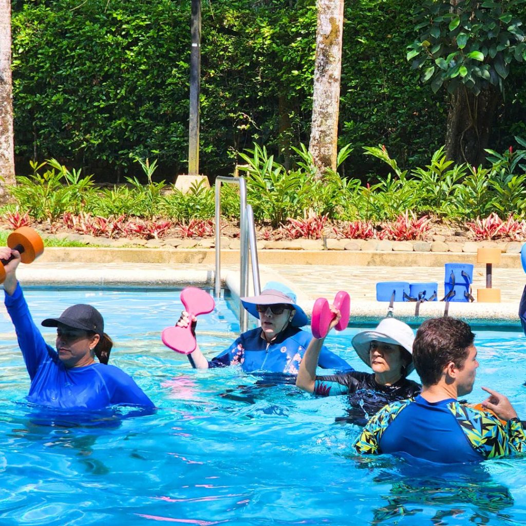 Grupo de personas y adultos mayores realizando ejercicios de AquaFitness con mancuernas de espuma en una piscina residencial. Instructora de AUA FIT dirigiendo una clase de acondicionamiento acuático de bajo impacto a domicilio en Cali.