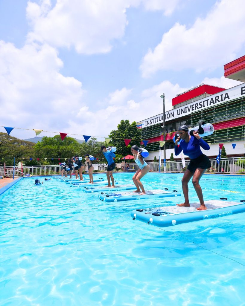Un grupo de personas en una piscina realizando sentadillas sobre tablas AquaBoard mientras sostienen waterbags sobre los hombros, durante una clase de AquaBoard Power.