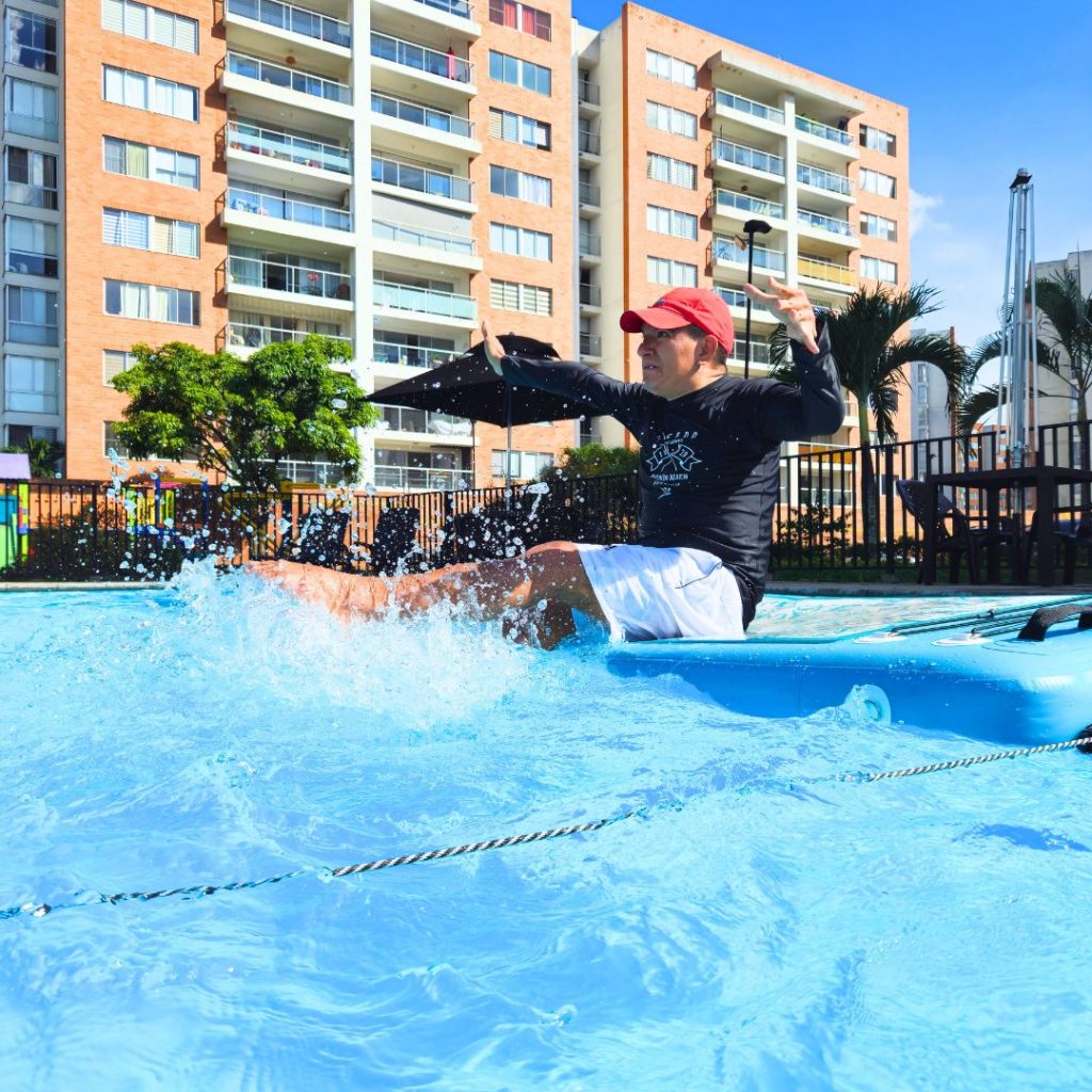Hombre con parálisis cerebral sonriendo y salpicando agua lleno de vitalidad sobre una tabla de AquaBOARD. Representación de la visión de AUA FIT: el agua como medio inclusivo para sanar, fortalecer y transformar vidas en Cali.